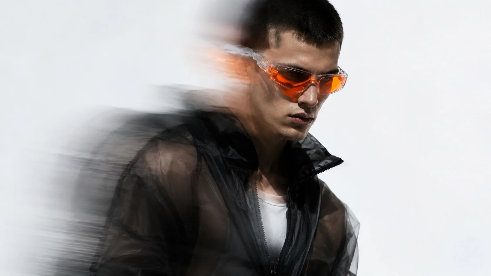 A model in a sheer black jacket and orange-tinted sunglasses against a bright white background, caught in motion blur